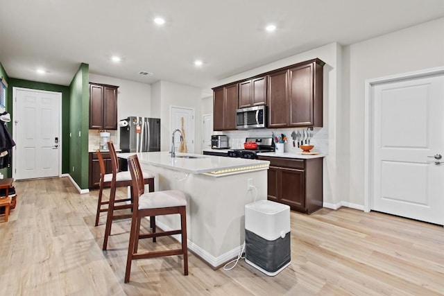 kitchen with sink, dark brown cabinets, a center island with sink, light wood-type flooring, and appliances with stainless steel finishes