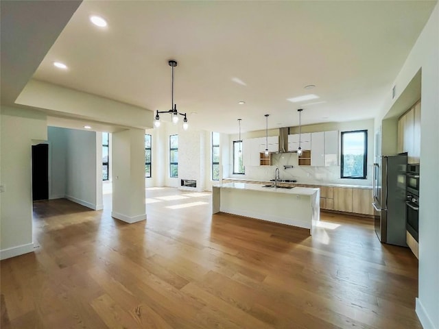 kitchen with stainless steel refrigerator, a kitchen island with sink, white cabinetry, ventilation hood, and decorative light fixtures