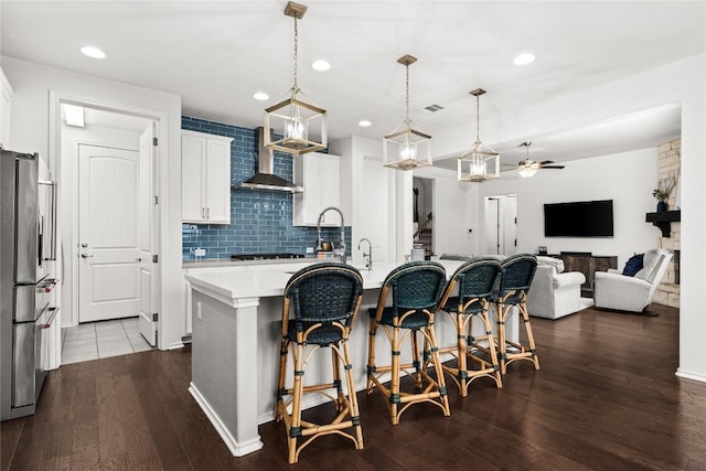 kitchen with appliances with stainless steel finishes, a kitchen island with sink, hanging light fixtures, white cabinets, and wall chimney exhaust hood