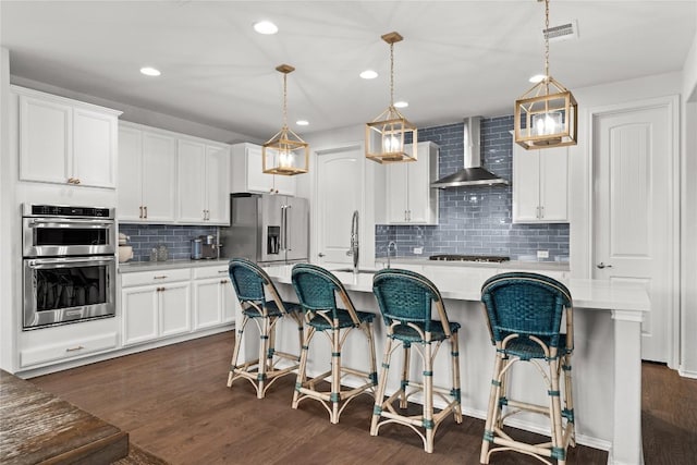 kitchen with pendant lighting, a kitchen island with sink, wall chimney range hood, and appliances with stainless steel finishes