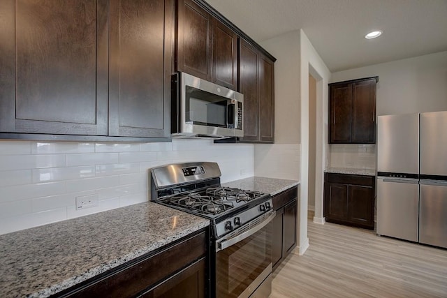 kitchen featuring light stone countertops, light wood-type flooring, stainless steel appliances, dark brown cabinets, and tasteful backsplash