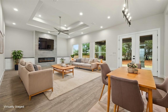 living room featuring ceiling fan, a raised ceiling, a tiled fireplace, french doors, and light wood-type flooring