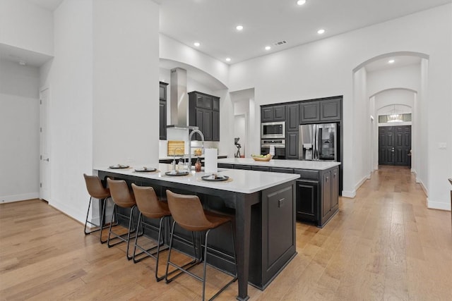 kitchen featuring an island with sink, a breakfast bar area, stainless steel appliances, wall chimney range hood, and light wood-type flooring