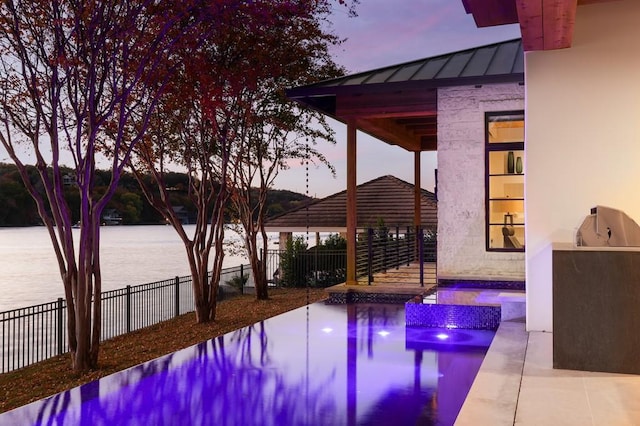 pool at dusk with area for grilling, a gazebo, a patio area, and a water view