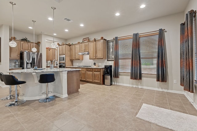 kitchen with appliances with stainless steel finishes, a breakfast bar, an island with sink, hanging light fixtures, and light stone counters