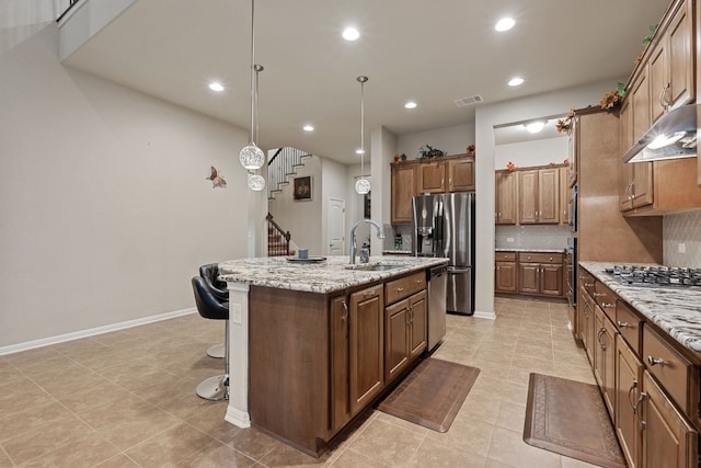 kitchen featuring sink, light stone counters, hanging light fixtures, appliances with stainless steel finishes, and an island with sink