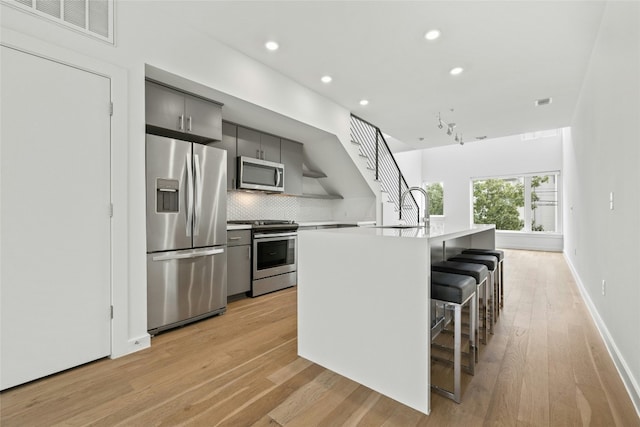 kitchen featuring gray cabinetry, backsplash, stainless steel appliances, a kitchen breakfast bar, and an island with sink