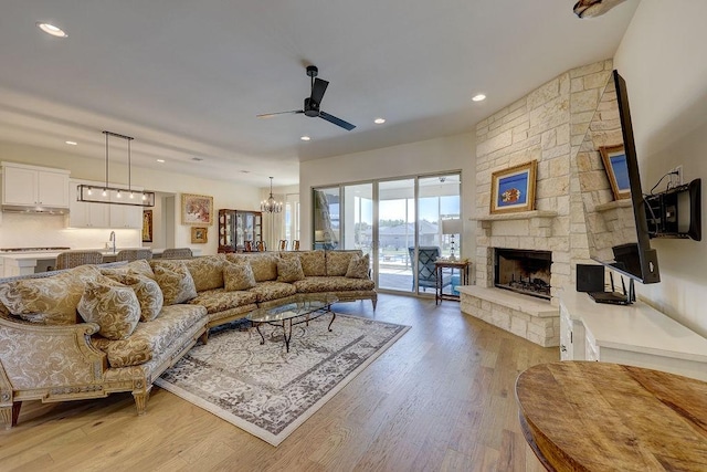 living room with a stone fireplace, ceiling fan with notable chandelier, and light wood-type flooring