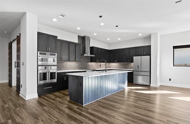 kitchen with dark wood-type flooring, a center island with sink, refrigerator with ice dispenser, a barn door, and wall chimney range hood