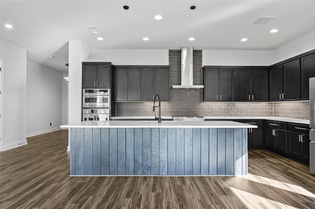 kitchen with wood-type flooring, sink, stainless steel appliances, a center island with sink, and wall chimney exhaust hood