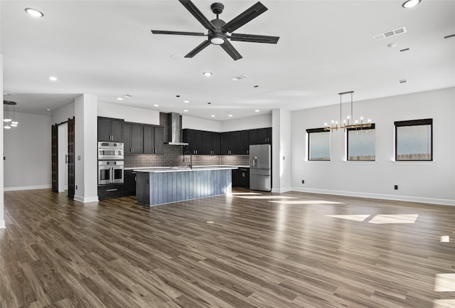 kitchen with appliances with stainless steel finishes, dark hardwood / wood-style flooring, a barn door, a kitchen island with sink, and wall chimney range hood