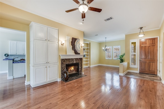 living room with crown molding, built in features, a fireplace, light hardwood / wood-style floors, and ceiling fan with notable chandelier