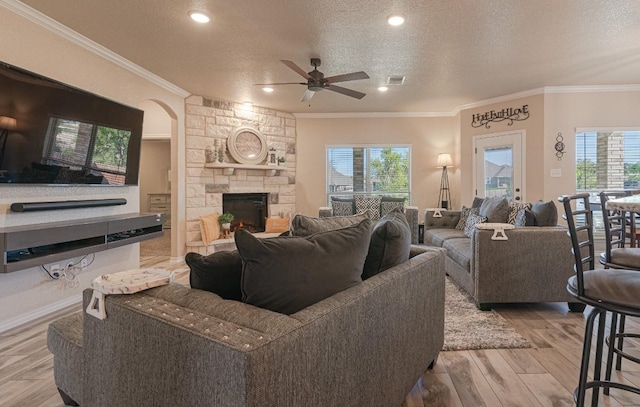 living room with a stone fireplace, crown molding, a textured ceiling, ceiling fan, and light hardwood / wood-style floors