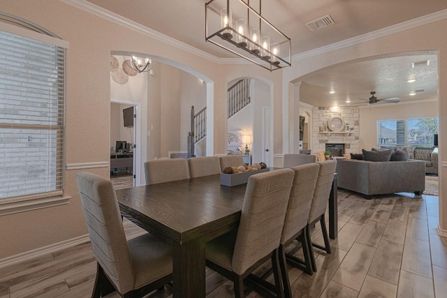 dining room featuring crown molding, a stone fireplace, ceiling fan with notable chandelier, and light hardwood / wood-style floors