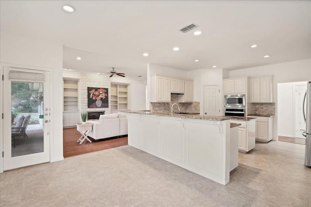 kitchen featuring appliances with stainless steel finishes, a large fireplace, ceiling fan, light stone countertops, and a kitchen island with sink