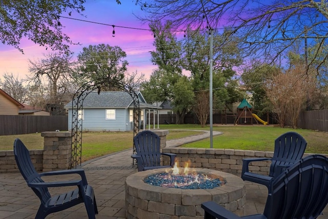 patio terrace at dusk with a playground, a yard, an outbuilding, and a fire pit