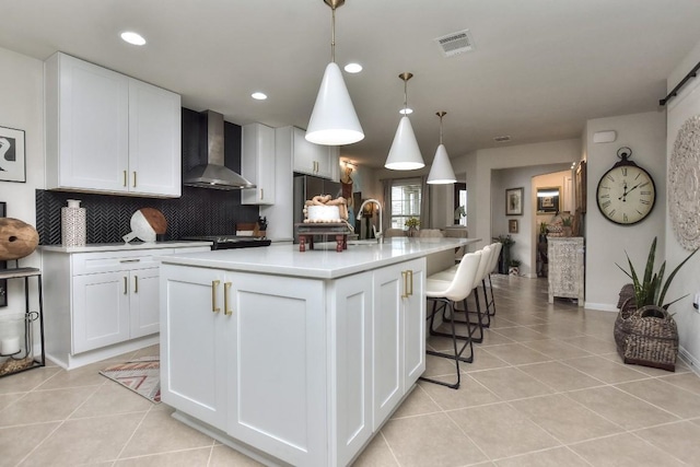 kitchen with pendant lighting, an island with sink, white cabinets, light tile patterned floors, and wall chimney range hood