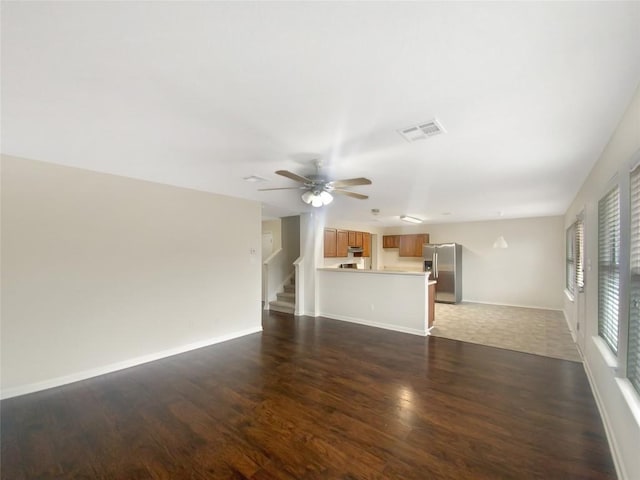 unfurnished living room featuring a ceiling fan, visible vents, baseboards, stairway, and dark wood-style floors