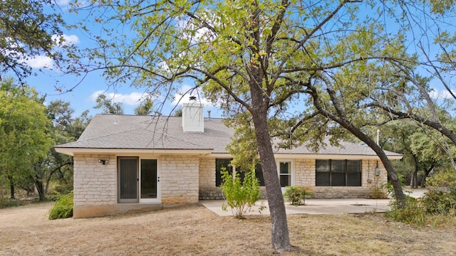 rear view of house featuring a patio area, a chimney, stone siding, and roof with shingles