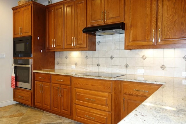 kitchen with brown cabinets, black appliances, under cabinet range hood, decorative backsplash, and light stone countertops