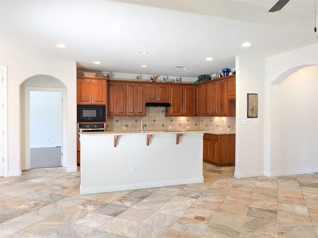 kitchen with arched walkways, under cabinet range hood, black microwave, brown cabinets, and backsplash