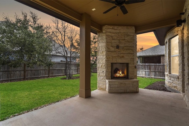 patio terrace at dusk with ceiling fan, a yard, and an outdoor stone fireplace