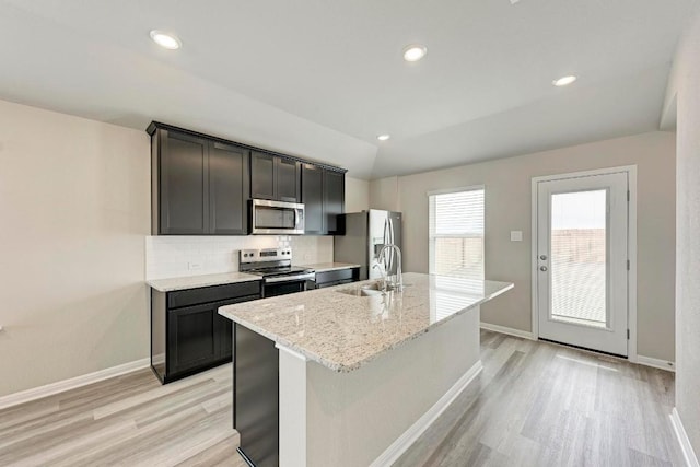 kitchen with appliances with stainless steel finishes, sink, backsplash, a kitchen island with sink, and light stone countertops