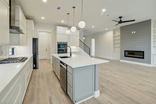 kitchen with wall chimney exhaust hood, an island with sink, black appliances, decorative light fixtures, and white cabinetry