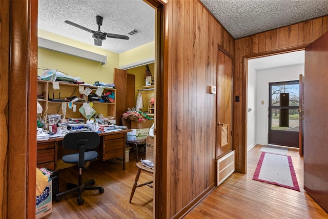 home office featuring ceiling fan, light hardwood / wood-style flooring, a textured ceiling, and wooden walls