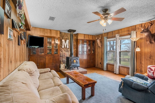 living room with a textured ceiling, ceiling fan, light hardwood / wood-style floors, wood walls, and a wood stove