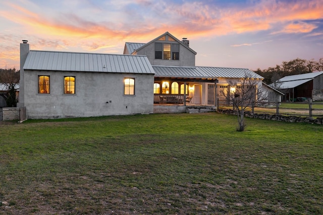 back of house at dusk featuring metal roof, a yard, stucco siding, a standing seam roof, and a chimney