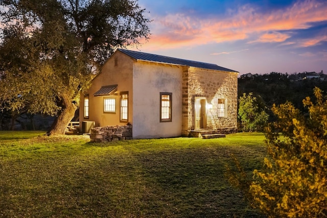 back of house at dusk featuring metal roof, stone siding, a yard, and stucco siding