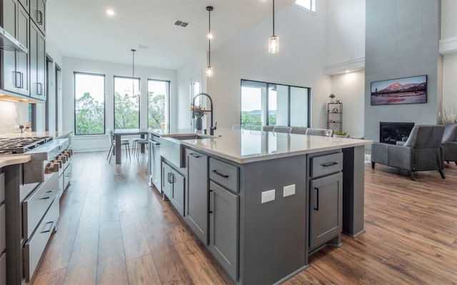 kitchen featuring a kitchen island with sink, gray cabinetry, a large fireplace, wall chimney exhaust hood, and pendant lighting