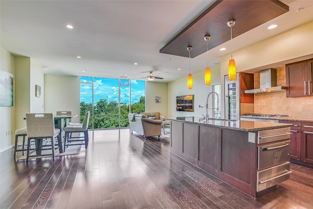 kitchen with sink, dark stone counters, wall chimney range hood, a wall of windows, and a kitchen island with sink