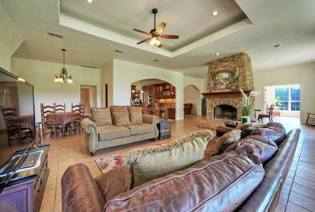 living room featuring a tray ceiling, a stone fireplace, and ceiling fan with notable chandelier