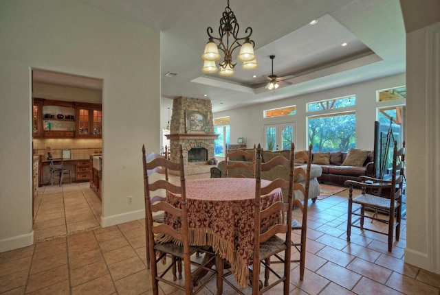 dining space featuring ceiling fan with notable chandelier, light tile patterned flooring, a stone fireplace, and a raised ceiling