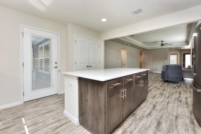 kitchen with dark brown cabinetry, stainless steel refrigerator, a tray ceiling, a kitchen island, and light wood-type flooring