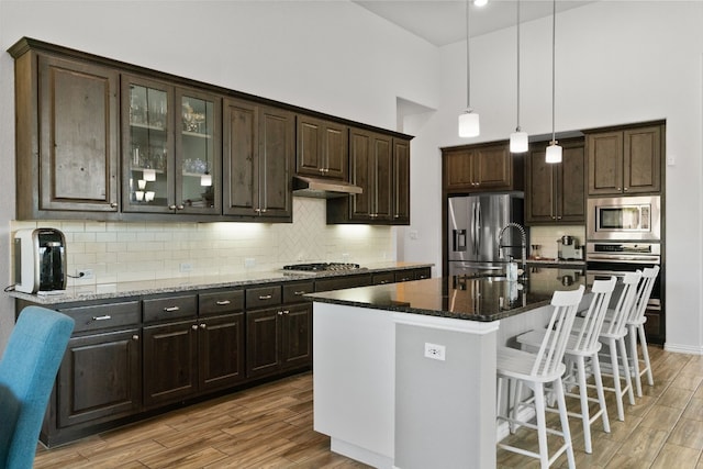 kitchen featuring stainless steel appliances, dark brown cabinetry, dark stone counters, decorative light fixtures, and a kitchen island with sink