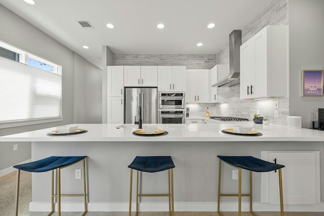 kitchen featuring white cabinetry, kitchen peninsula, appliances with stainless steel finishes, a kitchen breakfast bar, and wall chimney exhaust hood