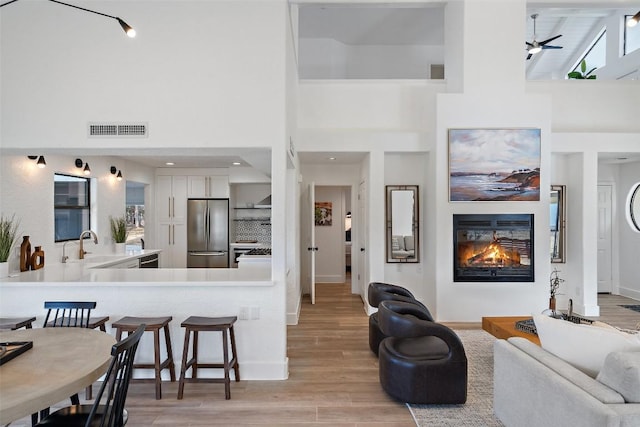 living room featuring a ceiling fan, visible vents, a towering ceiling, light wood-type flooring, and a glass covered fireplace