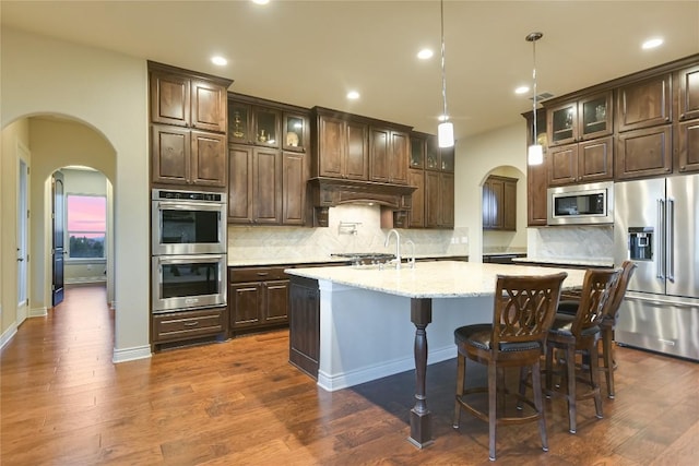 205 Palazza Alto Dr, Austin, TX 78734 - kitchen featuring appliances with stainless steel finishes, arched walkways, dark brown cabinetry, a kitchen island with sink, and recessed lighting