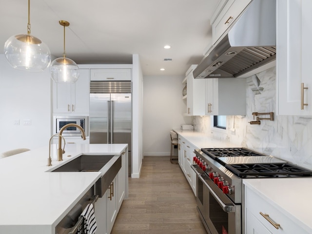 kitchen with built in appliances, hanging light fixtures, range hood, light countertops, and white cabinetry