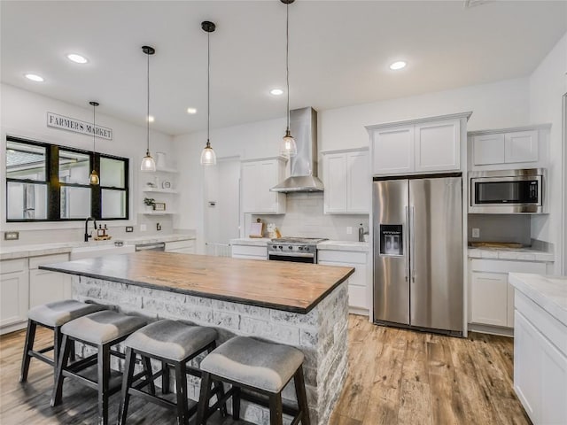 kitchen featuring wall chimney exhaust hood, stainless steel appliances, hanging light fixtures, white cabinetry, and a kitchen breakfast bar