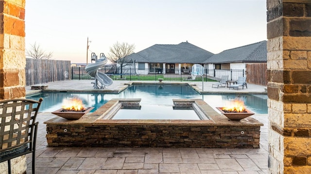 pool at dusk featuring a fire pit, a water slide, a fenced backyard, and a fenced in pool