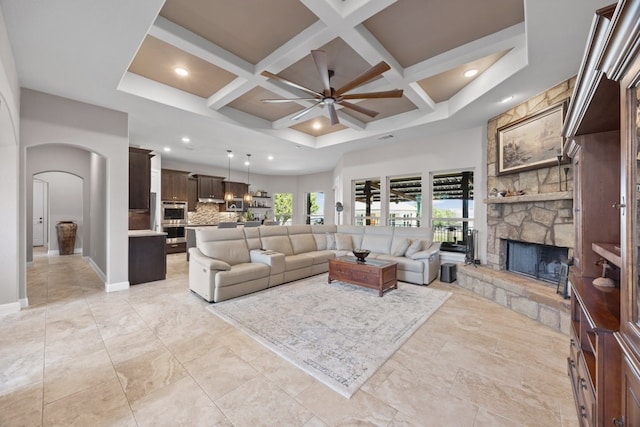 living area with arched walkways, a stone fireplace, coffered ceiling, a ceiling fan, and baseboards