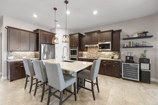 kitchen featuring beverage cooler, a center island with sink, appliances with stainless steel finishes, light countertops, and pendant lighting