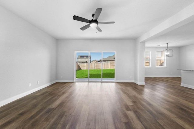 unfurnished living room with ceiling fan with notable chandelier, dark wood-type flooring, and baseboards