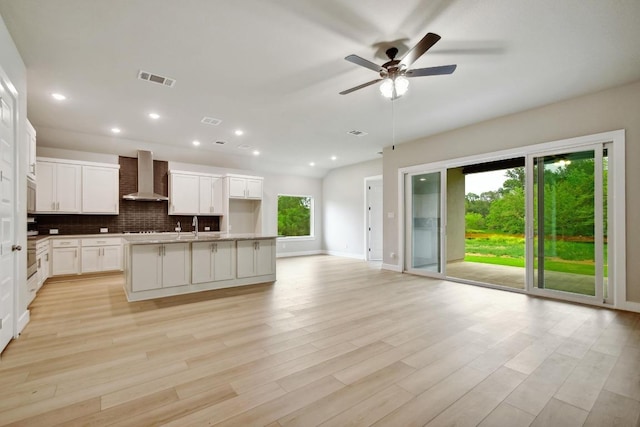 kitchen featuring open floor plan, a center island with sink, wall chimney range hood, a ceiling fan, and backsplash