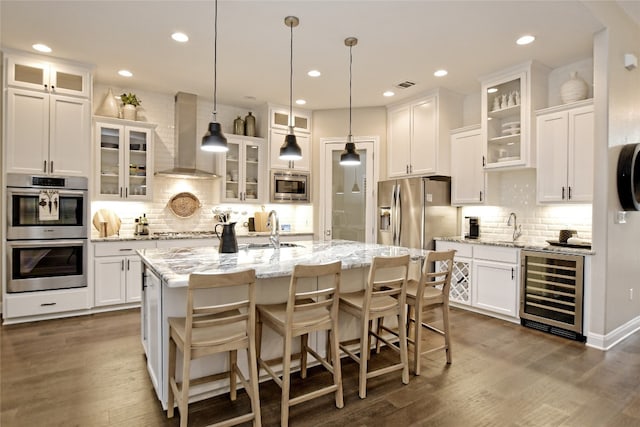 kitchen with beverage cooler, a center island with sink, wall chimney exhaust hood, glass insert cabinets, and appliances with stainless steel finishes