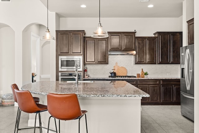 kitchen featuring stainless steel appliances, pendant lighting, a center island with sink, and light stone countertops